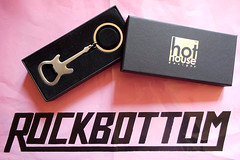 A brushed-metal bottle opener in the shape of an electric guitar, displayed in a black box with a lid reading “hot house designs”.  Part of a pink carrier bag with the logo “ROCKBOTTOM” is visible in the background.