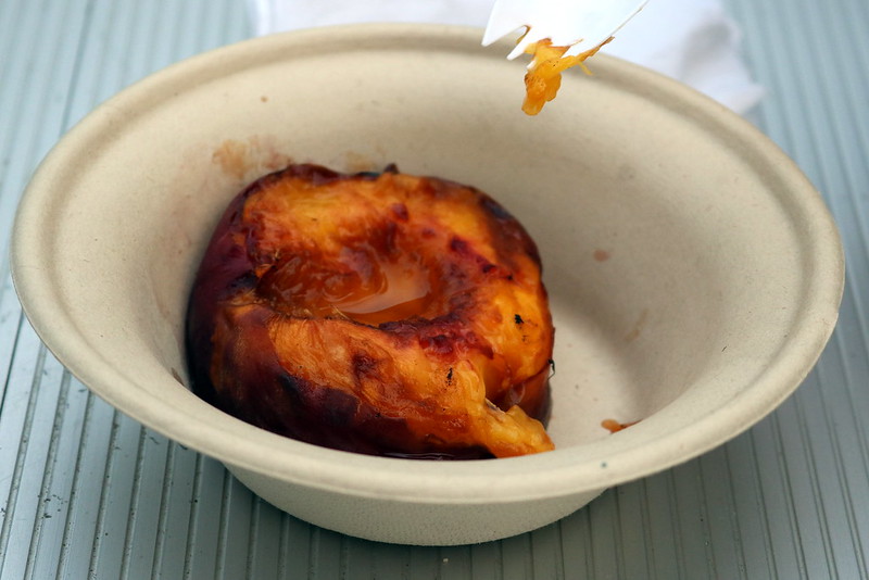 Half of a grilled peach with liquid in a compostable bowl.