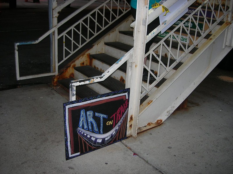 Rusty elevated staircase with painted "Art on Track" sign on ground at bottom