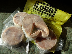 Four shrink-wrapped pieces of fish; three of them round and the other with a v-shape taken out of it.  The packets are resting on a yellow carrier bag with “Lobo Seafood Stores” printed on it.