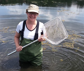A woman smiles while holding a net and wearing waders in water past her knees.