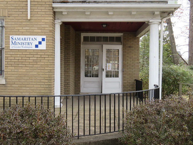 Photo of a yellow-brick building with white trim, a curving porch with a wrought-iron railing, and signage that reads 