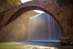 Waterfall beneath a very old arched bridge at Palaeokaryá, near Trikala, Greece.  Rays of hard sunlight paint the space below the bridge's arch into a dreamy, golden-coloured atmosphere despite the winter weather. The clear waters of Portaïkós river can inspire as well as cleanse…  The scenery reminds us of a poem written by the 17th century poet:  “With what deep murmurs through time's silent stealth Doth thy transparent, cool, and wat'ry wealth Here flowing fall, And chide, and call, As if his liquid, loose retinue stay'd Ling'ring, and were of this steep place afraid; … … As this loud brook's incessant fall In streaming rings restagnates all, Which reach by course the bank, and then Are no more seen, just so pass men…”  —Henry Vaughan (The Waterfall) 