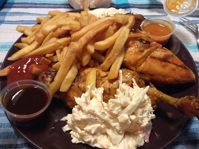 A large plate filled with thin, floppy chips, a grilled half-chicken on the bone, a couple of small plastic sauce pots, and a large dollop of coleslaw.
